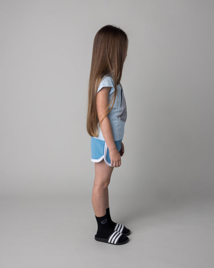 Kid wearing the Flower Penguin Tee in light blue with the Track Star Short in ocean, side view
